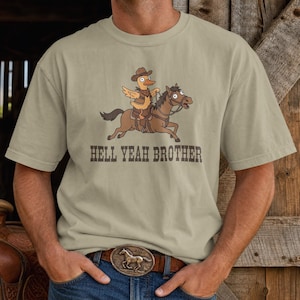 Puede incluir: Una camiseta beige de manga corta con un gráfico de un pato de dibujos animados con sombrero de vaquero y chaleco montado en un caballo marrón. Debajo de la ilustración, el texto HELL YEAH BROTHER está impreso en una fuente oscura y desgastada.