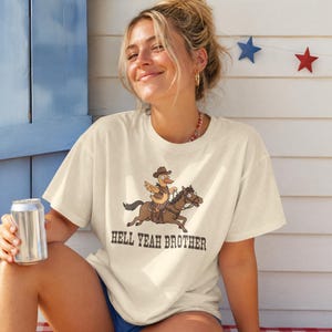 May include: A cream-colored short-sleeve t-shirt featuring a graphic of a cartoon duck wearing a cowboy hat and vest while riding a brown horse. Below the illustration, the text HELL YEAH BROTHER is printed in a dark brown, western-style font.
