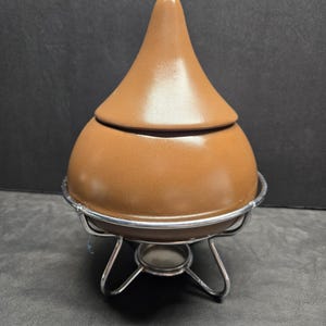 May include: A brown ceramic fondue pot with a lid, resting on a silver-colored metal stand. The pot has a rounded base and a conical lid. The stand has a circular base with three legs.