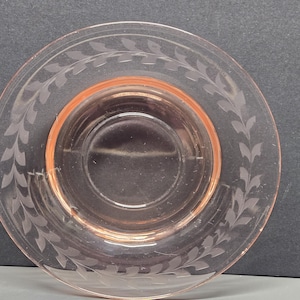 May include: A close-up of a pink glass saucer with a decorative etched leaf pattern around the rim. The saucer has a slightly raised center and a delicate, translucent appearance. The glass has a soft, rosy hue.