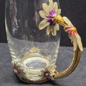 May include: Clear glass mug with a decorative handle and base. The handle and base are adorned with gold-colored metal and flower designs with purple and red accents. The mug has a wide, cylindrical shape.