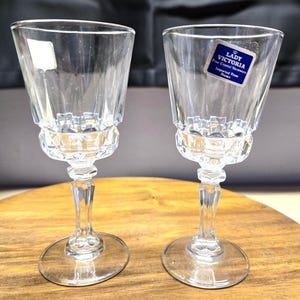 May include: Two clear crystal wine glasses with a faceted design. Each glass has a round base, a slender stem, and a flared bowl. One glass has a blue and white label that reads "LADY VICTORIA Fine Crystal Stemware Imported From France."