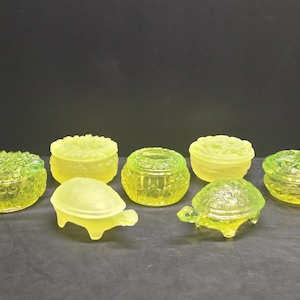 May include: A collection of yellow-green glass trinket boxes and turtle figurines. The boxes are round with lids, featuring floral and textured designs. The turtle figurines have detailed shell patterns and legs.