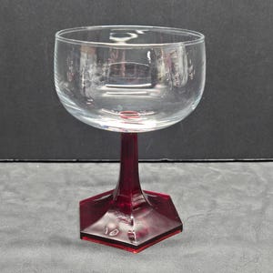 May include: A clear glass wine goblet with a burgundy stem and base. The bowl is round, and the stem is hexagonal. The goblet is designed for serving wine.
