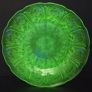 May include: A round, translucent neon green glass plate featuring a scalloped edge and a molded floral pattern. The design consists of raised cherry blossom motifs arranged in panels separated by vertical ridges, radiating from the center.