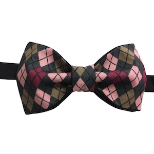 May include: A bow tie featuring an argyle pattern in shades of pink, dark gray, and tan. The fabric has a visible woven texture, and the design is accented with thin black lines forming a diamond grid. A black strap extends from both sides.
