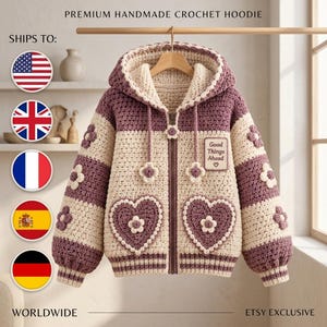 May include: A handmade crochet zip-up hoodie featuring alternating horizontal stripes of plum purple and cream yarn. The garment includes two large heart-shaped pockets, floral appliqués, and a patch with the text Good Things Ahead.