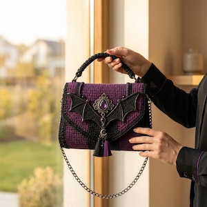 Pode incluir: Uma bolsa retangular de crochê em fio roxo escuro e preto. A aba apresenta um aplique preto em forma de morcego, um pingente central de metal ornamentado com uma gema roxa em forma de gota e um berloque de lua crescente com duas borlas.