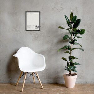May include: A framed rectangular wall art print featuring a white background with black text and a small black and white graphic element at the bottom, enclosed in a thin black frame, displayed on a textured gray wall above a white chair.