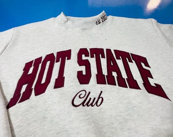 May include: A light gray heathered sweatshirt featuring the dark maroon text HOT STATE in a large arched font, with the word Club in a smaller script font below and the text est. 2027 embroidered on the right side of the mock neck collar.