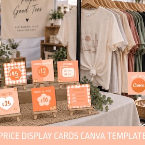 May include: A collection of six small square display cards on wooden easels, each featuring orange and white designs with text for sweaters, scented candles, stickers, t-shirts, mugs, and jewellery, displayed on a table with t-shirts on a rack.