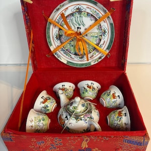 May include: A tea set featuring a teapot and six small cups with colorful floral and figure patterns, presented in a red fabric-covered box. A matching saucer is secured to the inside lid with orange ribbon, and the teapot is tied with string.