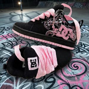 May include: A pair of black and pink DC skate sneakers featuring thick pink laces, a prominent DC logo on the side and tongue, and a silver cross charm attached to the eyelets. The design includes intricate white and pink scrollwork patterns.