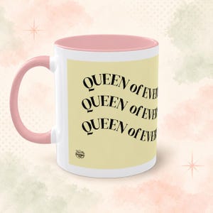 Könnte beinhalten: Ein weißer Keramikbecher mit einem zartrosa Henkel und Innenrand. Die Außenseite zeigt ein hellgelbes rechteckiges Feld mit dem schwarzen Text "QUEEN of EVERYTHING", der dreimal in einem wellenförmigen Design angeordnet ist.