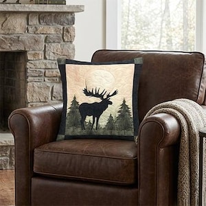 Peut inclure: Un coussin carré avec un fond matelassé de couleur crème, orné d'une grande silhouette sombre d'élan debout dans une forêt de pins sous une pleine lune. Le coussin est encadré par une bordure gris anthracite foncé sur les quatre côtés.