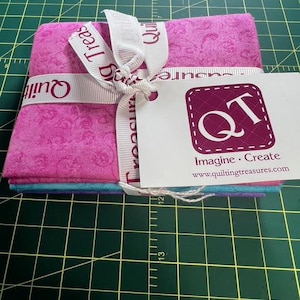 May include: A stack of pink and blue fabric squares tied with a white ribbon. The ribbon is printed with the words "Quilt Treasures". A white tag with the logo "QT" and the words "Imagine Create" is attached.