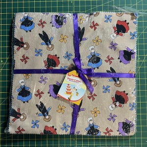 May include: A bundle of fabric squares wrapped in clear plastic and tied with a purple ribbon. The fabric features a repeating pattern of figures in dresses and bows. A tag with the words "Patty Cakes" is attached to the ribbon.