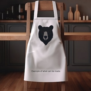 Puede incluir: Delantal de cocina blanco con un diseño de cara de oso negro. El texto "Ejemplo de lo que se puede hacer." está impreso en el delantal.
