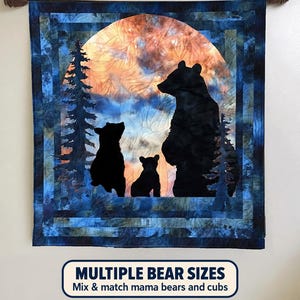 以下が含まれることがあります： 「Spindles and Quilts Grizzly 399 and Cubs Patterns」と題された正方形の布製壁掛け。オレンジと青の円形の夕日を背景に、大きなクマと2頭の子グマの黒いシルエットが描かれ、周囲には松の木と青い縁取りが施されています。
