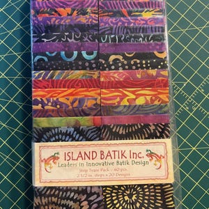 May include: A package of Island Batik fabric strips. The strips are 2 1/2 inches wide and feature a variety of colorful batik designs in shades of purple, orange, and black. The package includes 40 pieces and is labeled "Strip Tease Pack".