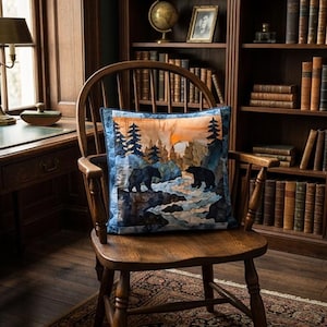 Può includere: Un cuscino quadrato decorativo con una stampa paesaggistica raffigurante due orsi neri vicino a una riva rocciosa. Lo sfondo mostra una foresta di conifere sotto un cielo al tramonto con tonalità arancioni e blu, rifinito da un bordo blu.