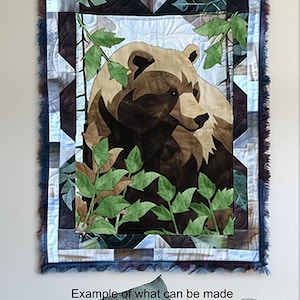 Może przedstawiać: Brązowy niedźwiedź z zielonymi liśćmi w tle. Niedźwiedź jest oprawiony w niebiesko-białą patchworkową kołdrę z geometrycznym wzorem.