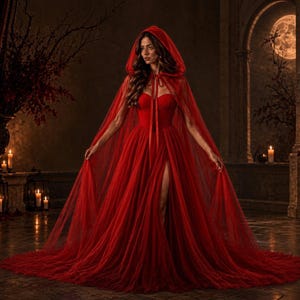 Pode incluir: Um vestido vermelho longo até ao chão com corpete sem alças e saia com fenda alta, combinado com uma capa vermelha transparente com capuz a condizer que se aperta no pescoço. O conjunto é feito de tecido de tule em camadas.