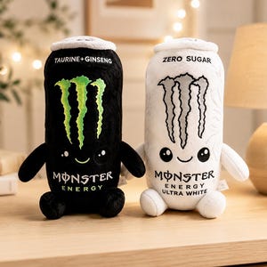 Pode incluir: Dois peluches em forma de latas de bebida energética. O peluche da esquerda é preto com um logótipo de garra verde, o texto TAURINE+GINSENG e MONSTER ENERGY. O da direita é branco com um logótipo de garra cinzento, o texto ZERO SUGAR e MONSTER ENERGY ULTRA WHITE.