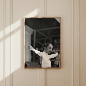 Op de afbeelding: Een zwart-wit ingelijste muurkunstprint met een artiest in een wit shirt met lange mouwen en een open rug, met grote oorringen, zingend in een microfoon met uitgestrekte armen op een podium met een structurele achtergrond.