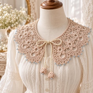 May include: A beige crochet collar featuring a floral pattern with scalloped edges and light blue beads along the perimeter. The collar includes a central twisted cord tie with two matching crochet tassels at the ends, worn over a white blouse.