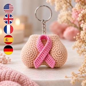 May include: A crochet keychain featuring a beige, rounded, textured form with a prominent pink ribbon stitched onto the center. The item is attached to a silver-toned metal split ring and a short chain. Five circular flag icons are arranged vertically on the left.