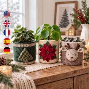 May include: Three cylindrical crochet plant pot covers. The left is dark green with a textured light green tree. The centre is cream with a red poinsettia flower. The right is light brown with a beige reindeer face featuring brown antlers.
