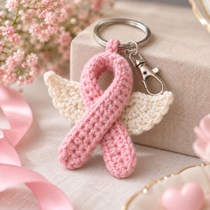 May include: A crochet keychain featuring a pink awareness ribbon with attached cream-colored wings. The ribbon is constructed from yarn and is connected to a silver-toned metal split ring and a lobster clasp for secure attachment to items.