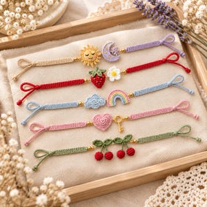 May include: Five handmade crochet bracelets featuring various charms: a yellow sun and purple moon, a red strawberry and white flower, a blue cloud and rainbow, a pink heart and gold key, and two green cherry stems with red cherries.