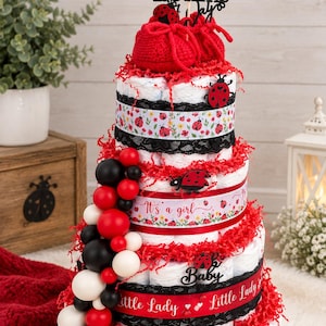 May include: A three-tiered diaper cake decorated with ladybug motifs, red and black ribbons, black lace, and red paper shred. It features a cake topper reading A Little Love Bug Is On The Way, red crocheted baby booties, and a balloon garland.