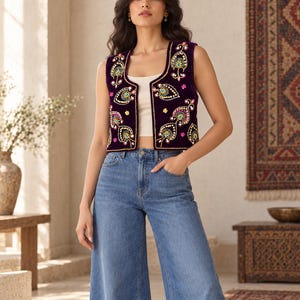 May include: A deep purple velvet waistcoat featuring intricate multi-coloured embroidery with floral and paisley motifs in shades of blue, green, yellow, and pink, paired with a simple cream-coloured tank top and wide-leg blue denim jeans.