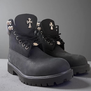 May include: A pair of black leather boots featuring silver-toned metal cross emblems on the tongues, silver eyelets, and decorative silver hardware accents along the collars. The boots have black laces and thick, lugged black rubber outsoles.