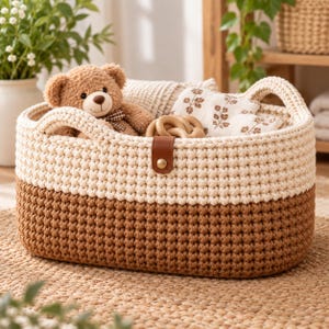 May include: An oval-shaped storage basket made of thick, hand-crocheted yarn with a two-tone design featuring a cream-colored top and a brown bottom. The basket includes two integrated handles and a brown faux leather tab with a gold metal snap.
