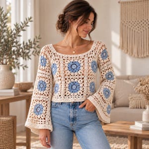 May include: A long-sleeved crochet top featuring a cream-colored base with a repeating pattern of light blue floral motifs. The garment has a wide scoop neckline and flared sleeves, constructed with an openwork stitch design throughout the body.