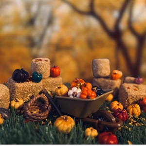 May include: A miniature brown wheelbarrow filled with small orange, white, and black pumpkins, surrounded by several square hay bales and scattered miniature pumpkins on a green surface, creating a harvest-themed display of tiny objects.