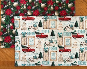 May include: A rectangular quilted placemat featuring a pattern of red trucks, wooden sleds, pine trees, and the text "NOEL" and "Walking in a Winter Wonderland" layered over a second placemat with a pattern of red poinsettias and silver ornaments.