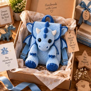 Puede incluir: Una mochila de dragón tejida a ganchillo en azul claro con detalles en azul oscuro, cuernos blancos y grandes ojos negros. La mochila está dentro de una caja de regalo de cartón con papel de seda y una etiqueta A Special Gift for a Special Kid.