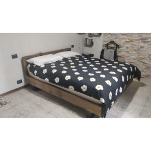 Könnte beinhalten: Ein Holzbettrahmen mit dunkelbraunem Finish und einem schwarzen Bettbezug, der mit einem sich wiederholenden Muster aus weißen und gelben Spiegeleiern bedruckt ist. Zwei weiße Kissen liegen am Kopfende des Bettes auf einer weißen Matratze.