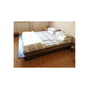 Op de afbeelding: Een laag platformbedframe van donkerbruin hout met een brede rand. Het bed is opgemaakt met een grijs-wit dekbedovertrek met patroon, een opgevouwen wit dekbed en twee witte kussens die tegen het hoofdeinde aan liggen.