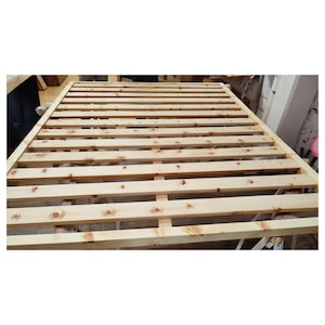 Op de afbeelding: Een rechthoekig bedframe opgebouwd uit lichtgekleurde natuurlijke houten latten. Het frame is voorzien van een reeks horizontaal geplaatste houten planken met gelijke tussenruimtes, ondersteund door een centrale verticale balk.