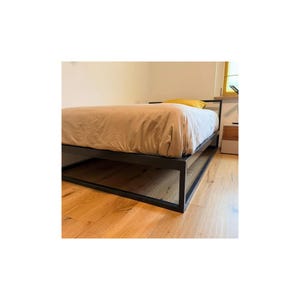 Könnte beinhalten: Ein niedriges Bettgestell aus schwarzem Metall mit rechteckigem Unterbau. Das Bett ist mit einem hellbeigen Bettbezug und einem einzelnen senfgelben Kissen am Kopfende vor einer schlichten weißen Wand bezogen.