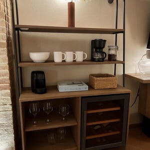 Peut inclure: Une étagère haute à structure métallique sombre et tablettes en bois. Elle comprend un bol blanc, trois tasses blanches, une cafetière noire, un bocal en verre, une machine à capsules noire, un livre, un panier tressé et des verres à vin.