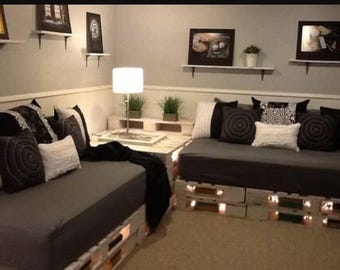 May include: Two low-profile sofas constructed from white-painted wooden pallets, topped with dark grey cushions. The seating area features an assortment of black, white, and grey throw pillows, a white table lamp, and small potted green plants.