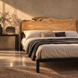 Puede incluir: Una estructura de cama con un cabecero de madera natural de borde irregular con vetas marcadas, sobre una base de metal negro. La cama está vestida con ropa de cama en color beige claro, una manta gris oscuro y dos cojines rectangulares.