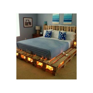 May include: A bed frame constructed from light-colored wooden shipping pallets with integrated warm yellow lighting within the base. The bed features a blue and white striped mattress cover and two blue and white patterned pillows.